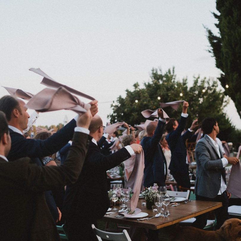die Gäste des Brautpaarstehen an großen Tischen in dem Garten eines Weingut in Barcelona, über ihnen hängen Glühbirnenlichterketten, die Sonne geht langsam unter, die Gäste schwingen über ihren Köpfen große Leinentücher