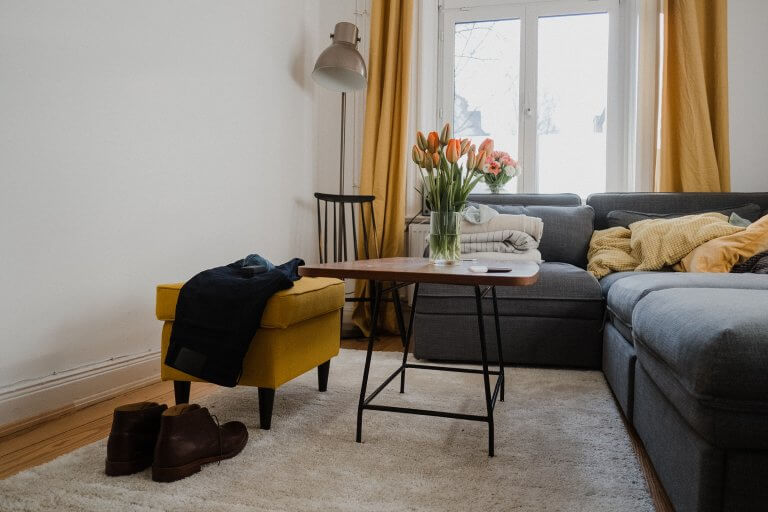 ein Wohnzimmer mit großen Sofa, es steht ein Tulpenstrauß auf dem kleinen Tisch, auf dem gelben Hocker liegt ein Sakko, davor stehen braune Schuhe
