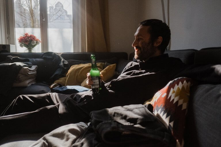 ein Mann sitzt auf dem Sofa, er hält ine Flasche Bier in der Hand, die Sonne scheint durch das Fenster