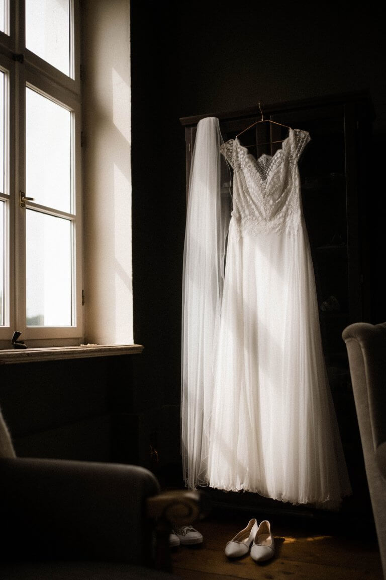 Hochzeitsfotografie La Dersentina. Das Brautkleid höngt an einem Kleiderbügel am Fenster in der Villa, der Schleier hängt daneben, die Brautschuhe stehen davor auf dem Boden, die Sonne scheint in das Fenster