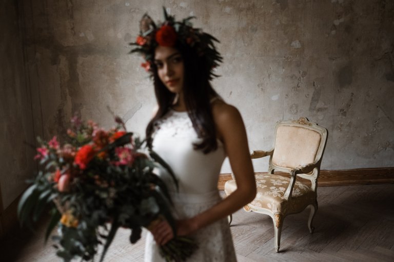 Brautmode Hamburg- AveevA- vor einer naturbelassenen Wand in Haller 6 steht ein antiker Sessel, im Vordergrund steht eine Frau in einem Brautkleid, sie hält einen üppigen Blumenstrauß in der Hand und sie trägt einen großen Blumenkranz auf dem Kopf