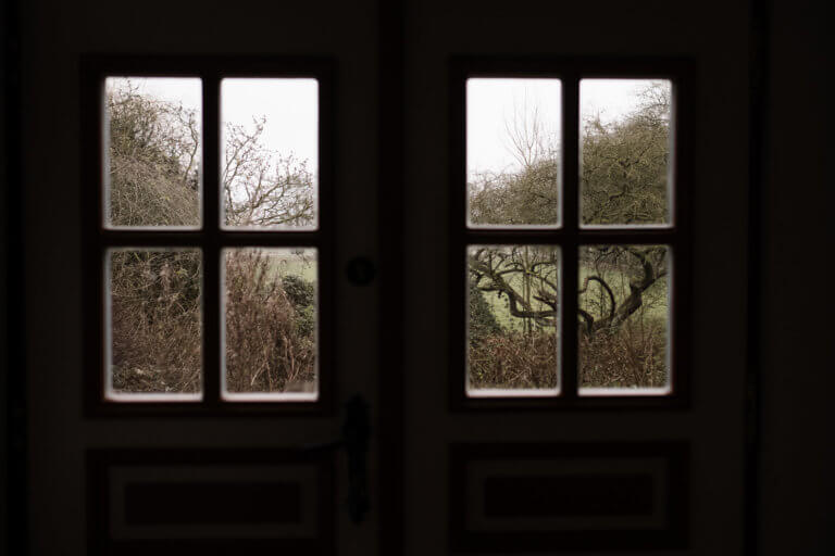der Blick aus einem großen, breiten Holzfenster mit Sprossen, der Blick ist auf kahle Bäume und einen kahlen Garten