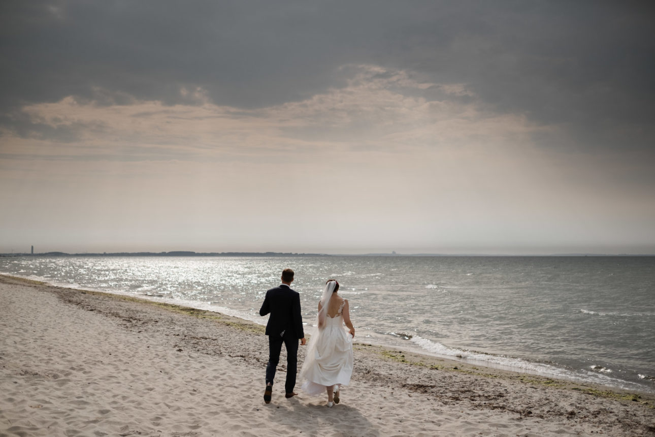 Schlossgut-Groß-Schwansee- Hochzeitsfotografie- Hochzeitsbilder an der Ostsee- Hochzeitspaar geht auf die See zu. Über ihnen ein dramatischer Himmel.